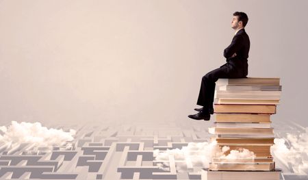 A serious businessman in suit sitting on a pile of giant books in front of a grey wall with clouds, labirynth