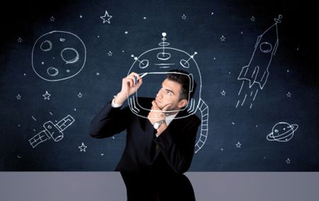 A young happy businessman in elegant suit drawing helmet, satellite and rocket in empty space with a chalk illustration concept