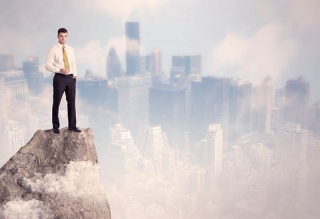 A successful good looking businessman standing on top of a high cliff above the city scape with clouds concept