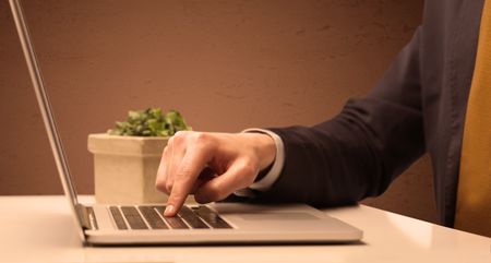 An office worker in elegant suit sitting at desk, typing on portable laptop with empty brown wall background