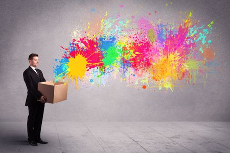 A young smiling business male holding a paperboard box with illustration of colourful spray paint splash on urban wall background concept.