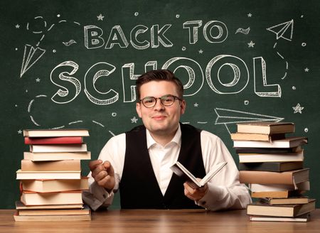 A young teacher in glasses sitting at classroom desk with pile of books in front of blackboard saying back to school drawing concept.