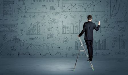 A businessman in modern stylish elegant suit standing on a small ladder and drawing pie and block charts on grey wall background with exponential progressing curves, lines, circles, angles, numbers