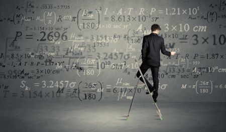 A businessman in elegant suit standing on a small ladder and writing numbers, calculating on grey wall background