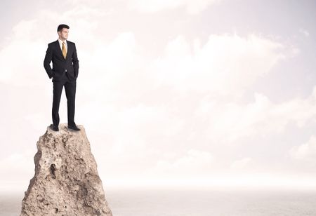 Successful sales person with brief case standing on top of a mountain cliff edge looking above the landscape between the clouds