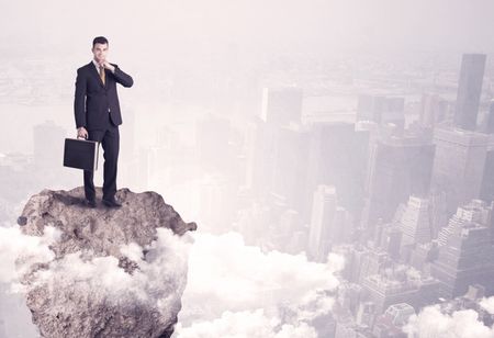 A young successful happy businessman standing confident on stone cliff in the clouds looking at the city scape from above concept