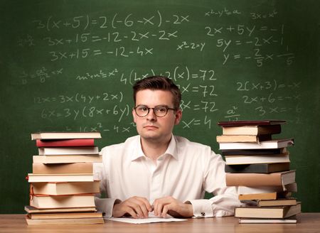 A young ambitious teacher in glasses sitting at classroom desk with pile of books in front of blackboard full of math calculations, numbers, back to school concept.