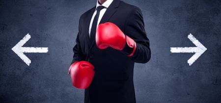 A confident businessman standing with red boxing gloves on his hand in front of arrows pointing in different directions on urban wall background concept.