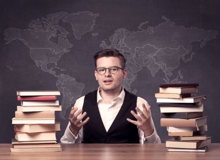 A young ambitious geography teacher in glasses sitting at classroom desk with pile of books in front of world map drawing on blackboard, back to school concept.