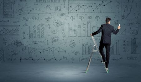 A businessman in modern stylish elegant suit standing on a small ladder and drawing pie and block charts on grey wall background with exponential progressing lines, circles, angles, blocks, numbers