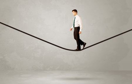 An elegan businessman in suit balancing on a tight rope with a briefcase in front of grey urban wall background environment concept