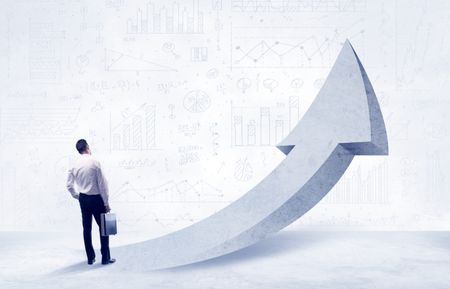 Young sales business person in elegant suit standing with his back in front of a big arrow pointing up and a clear background full of pie charts, numbers