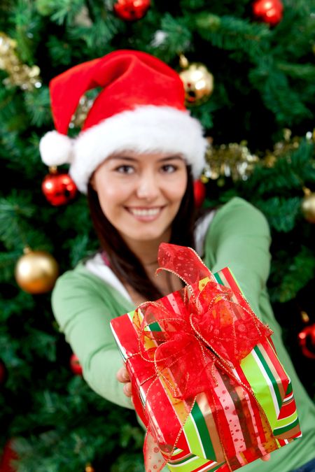 Woman next to the tree with a Christmas gift and wearing a Santa hat