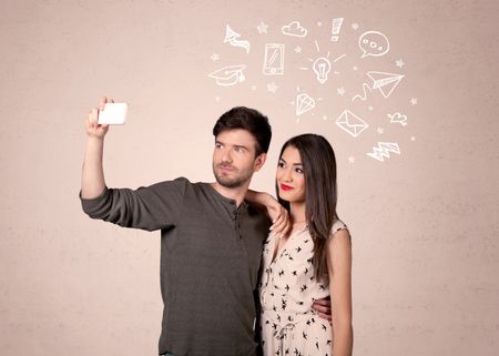 A young couple in love taking selfie with a mobile phone in the handsome guy's hand and drawn media communication icons above them, confused ideas concept