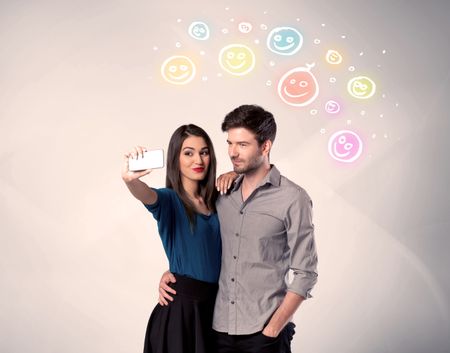 A cheerful young couple taking selfie photo with mobile phone and colorful happy smiley faces illustration above them concept