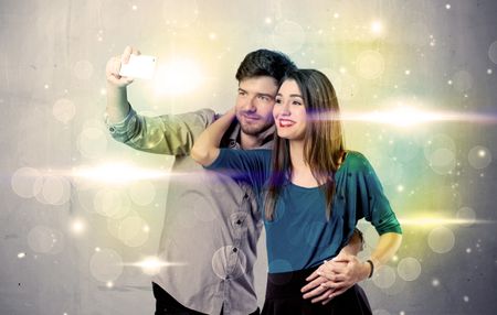 A fresh happy couple taking selfie photo with mobile phone in front of colorful lights glitter wall background concept