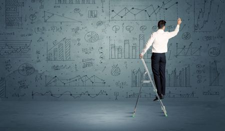 A businessman in modern stylish elegant suit standing on a small ladder and drawing pie and block charts on grey wall background with exponential progressing curves, lines, circles, blocks, numbers