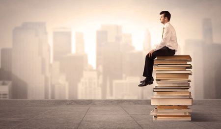 A serious student in elegant suit sitting on a pile of books looking over a brown sepia city landscape