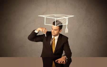 An elegant, successful university student drawing himself a square academic mortarboard cap with a chalk in front of grey wall background concept