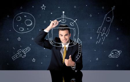 A young happy businessman in elegant suit drawing helmet, satellite and rocket in empty space with a chalk illustration concept