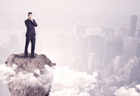 A young successful happy businessman standing confident on stone cliff in the clouds looking at the city scape from above concept