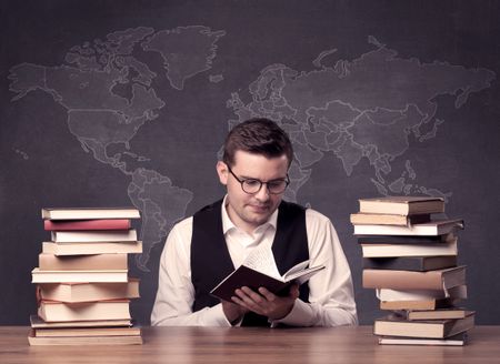 A young ambitious geography teacher in glasses sitting at classroom desk with pile of books in front of world map drawing on blackboard, back to school concept.