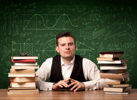 A young passionate male teacher sitting at school desk, reading a book, with area algorythm calculations and numbers on the blackboard concept.