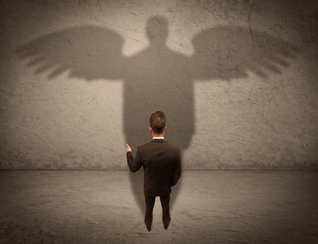 A successful businessman holding a briefcase is giving thumbs up for his angel shadow in front of a wall concept