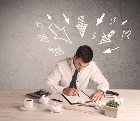 A young businessman sitting at an office desk and working on paperwork with drawn arrows pointing at his head concept 