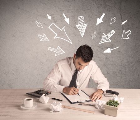 A young businessman sitting at an office desk and working on paperwork with drawn arrows pointing at his head concept