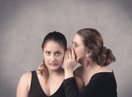 Two fancy dressed actress girls with long hair and make up whispering in front of grey urban background concept.