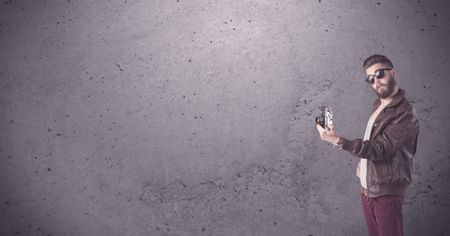 A stylish funny hipster person holding a vintage camera and taking photographs in front of a concrete clear empty urban wall background concept