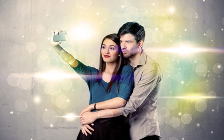 A fresh happy couple taking selfie photo with mobile phone in front of colorful lights glitter wall background concept