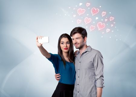A young couple in love and drawn red hearts taking selfie with a mobile phone in the handsome guy's hand in front of an empty clear grey wall background concept