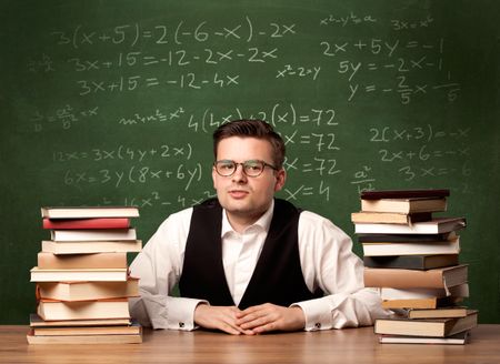 A young ambitious teacher in glasses sitting at classroom desk with pile of books in front of blackboard full of math calculations, numbers, back to school concept.