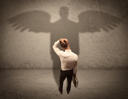 A successful businessman holding a briefcase is giving thumbs up for his angel shadow in front of a wall concept