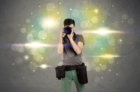 A young amateur photographer with professional camera equipment taking picture in front of grey wall full of colorful bokeh and glowing lights concept