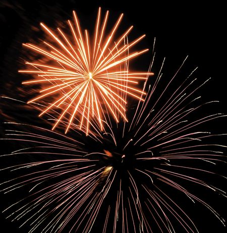 Two bursts of fireworks, the larger with silvery and pink streaks, the other yellow-white with reddish smoke