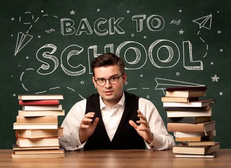 A young teacher in glasses sitting at classroom desk with pile of books in front of blackboard saying back to school drawing concept.