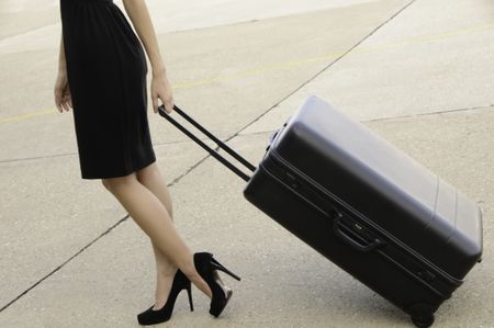 Young businesswoman in high heels and black dress drags a black suitcase across tarmac at airport