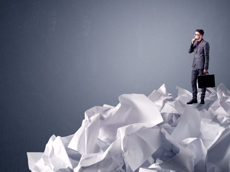 Thoughtful young businessman standing on a pile of crumpled paper with a light grey background