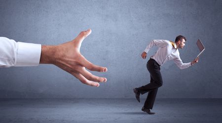 Young miniature businessman running from a big hand with blueish grey background