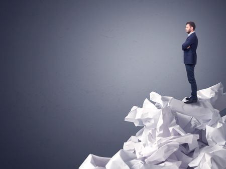 Thoughtful young businessman standing on a pile of crumpled paper with a dark grey background