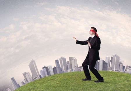 Young blindfolded businessman steps on a a patch of grass with a grey buildings in the background