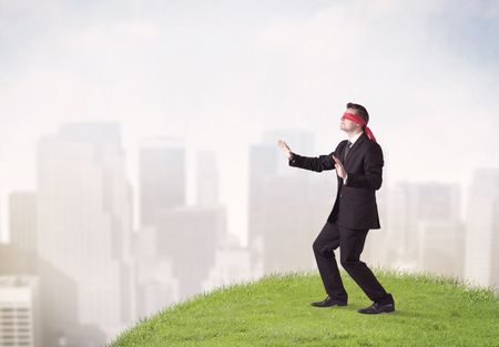 Young blindfolded businessman steps on a a patch of grass with a city in the background 