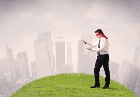 Young blindfolded businessman steps on a a patch of grass with a cloudy city in the background