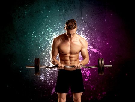 A sexy male fitness trainer showing his muscles and looking seductive with a weight in his hands in front of bright paint splash purple wall concept