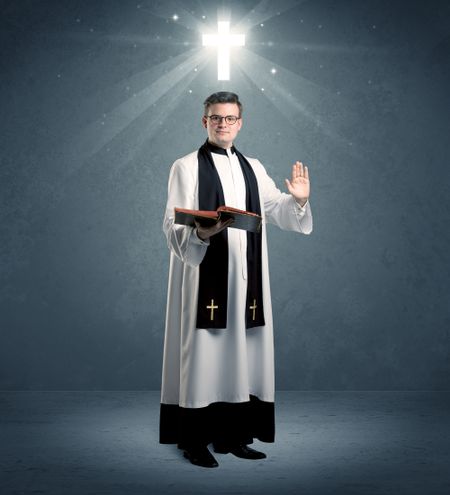 A young caucasian priest with deep faith blessing with the holy bible in his hand in front of blue wall background with illustrated glowing cross concept.