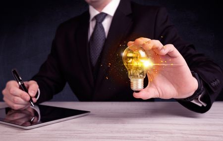 An elegant office worker holding a yellow sparkling light bulb in his hand while working in front of dark blue background concept.