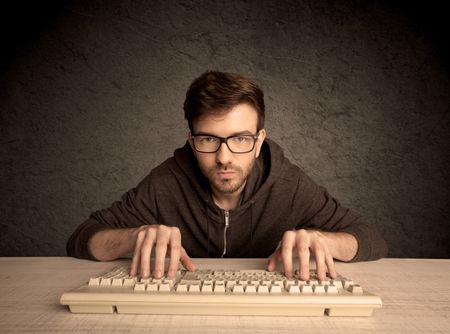 A young hacker with glasses dressed in casual clothes sitting at a desk and working on a computer keyboard in front of black clear concrete wall background concept